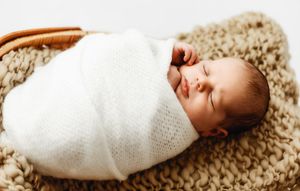 Peaceful newborn baby swaddled in white knit blanket sleeping in a wicker basket on a cozy brown blanket.