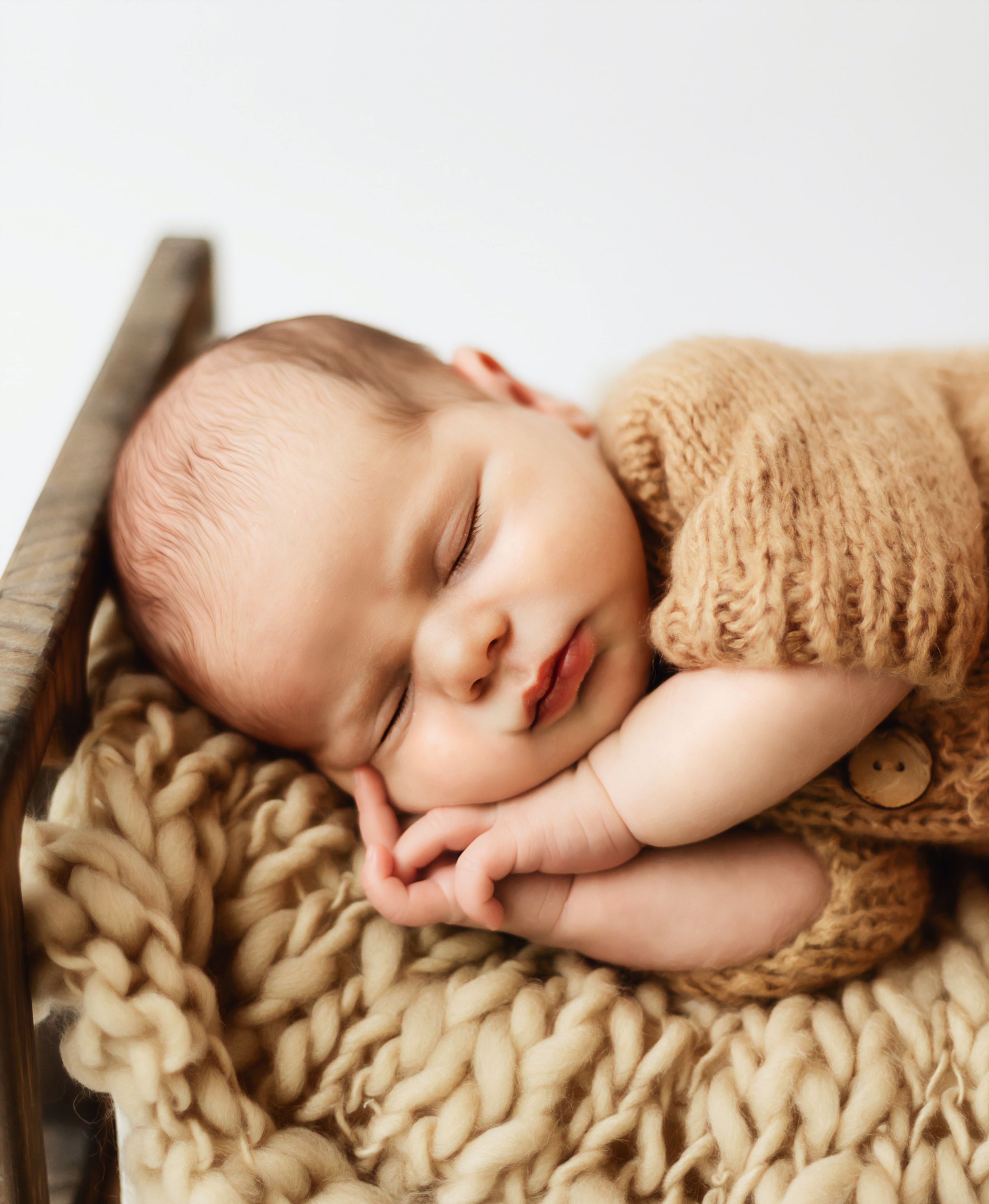 Peaceful newborn baby sleeping in a rustic wooden bed, wearing a cozy knit tan romper on a chunky wool blanket.