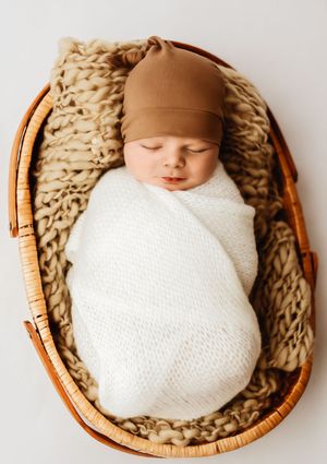 Sleeping newborn baby swaddled in white wrap wearing brown hat, resting in wicker basket with chunky knit blanket.