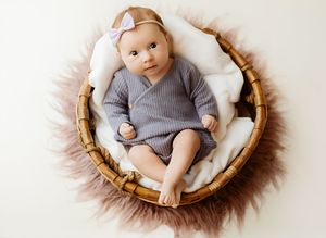 Newborn baby in gray romper and pink bow headband resting in a wicker basket with fluffy white fur lining.