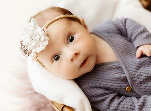Adorable baby girl wearing a floral headband and gray quilted romper, looking up with big brown eyes.