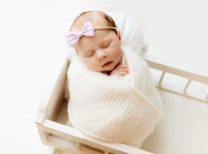 Sleeping newborn baby girl wrapped in white knit swaddle wearing pink bow headband in wooden mini bed.