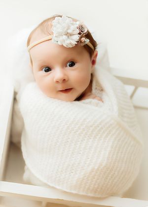 Adorable newborn baby girl swaddled in white knit wrap wearing floral headband, posed in white crib.