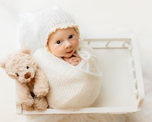 Newborn baby wrapped in white knit swaddle wearing a white hat, sitting in a white crate with a teddy bear.
