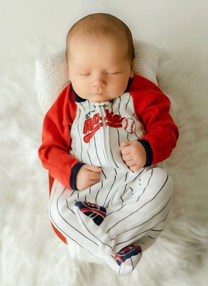 Sleeping newborn baby wearing a baseball-themed pinstripe onesie with red jacket, resting on a white fluffy blanket.