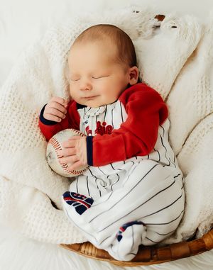 Sleeping newborn baby in baseball uniform onesie holding a baseball, nestled in a basket with white blanket.