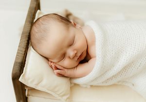 Peaceful newborn baby sleeping wrapped in white knit blanket on cream pillow in tiny wooden bed.