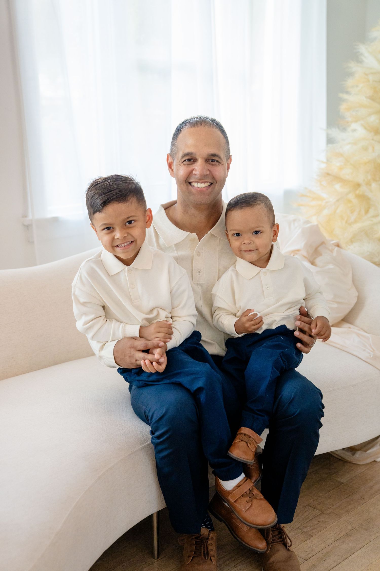Smiling father sits on white sofa with two young sons, all wearing matching white shirts and navy blue pants.