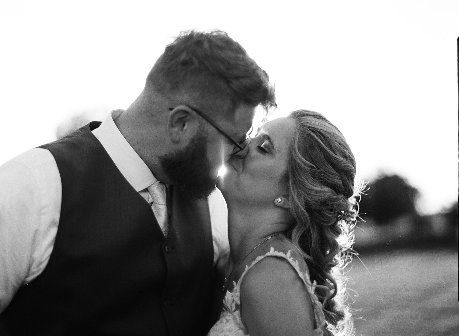 Black and white wedding photo of couple sharing a romantic kiss, bride in lace dress, groom in vest and glasses.