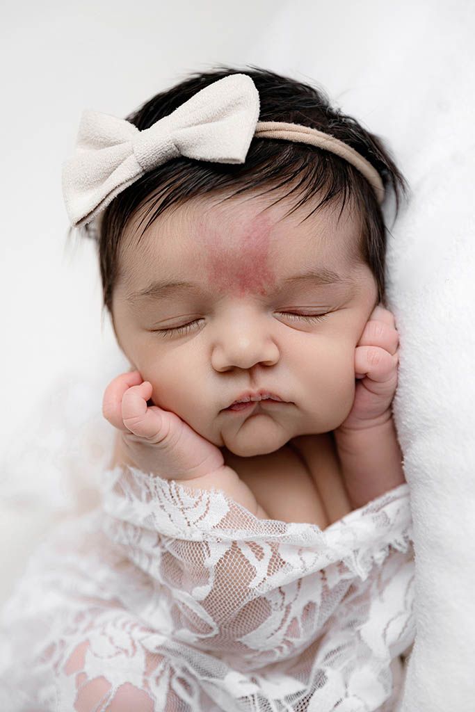 Peaceful newborn baby girl sleeping wrapped in white lace with a cream bow headband on white background.