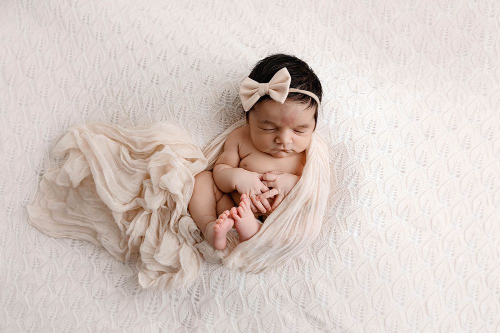 Sleeping newborn baby girl wrapped in cream fabric with bow headband on white background.