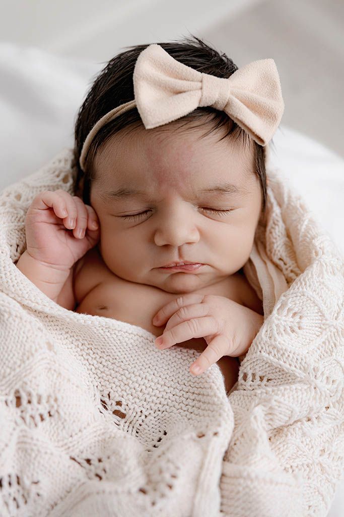 Sleeping newborn baby girl wrapped in a cream knit blanket wearing a beige bow headband.