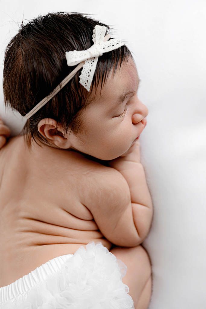 Sleeping newborn baby girl with dark hair wearing a white lace bow headband and ruffled diaper cover.
