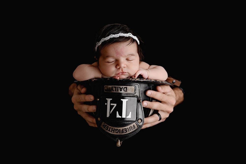 Sleeping newborn baby girl resting on a firefighter helmet badge, held by adult hands against black background.