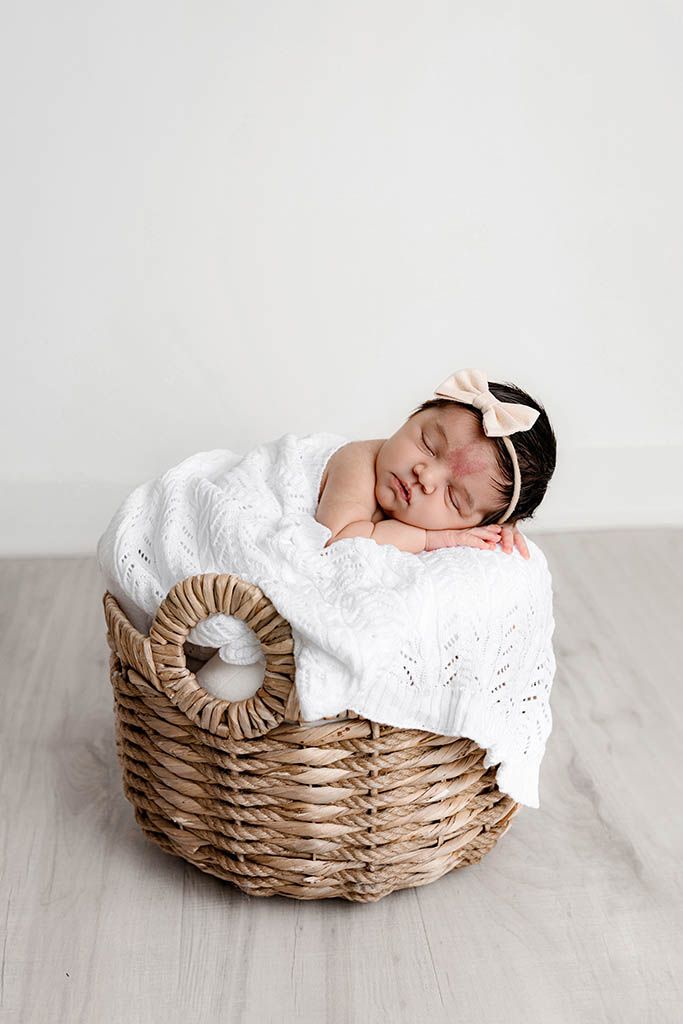 Sleeping newborn baby girl in a wicker basket wrapped in a white knit blanket with a pink bow headband.