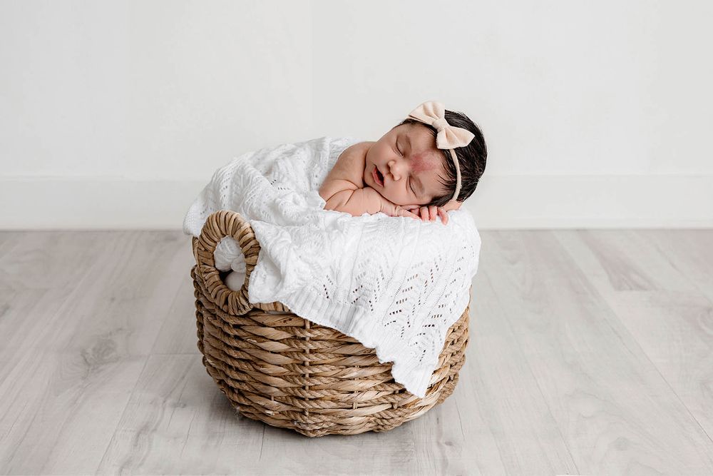 Newborn baby sleeping peacefully in a wicker basket with a white knit blanket and bow headband.