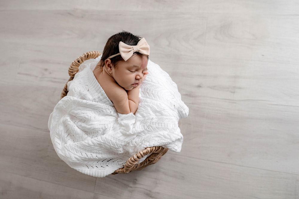 Sleeping newborn baby girl wrapped in white knit blanket with bow headband in wicker basket.