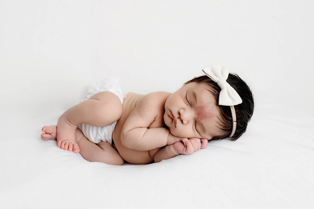 Sleeping newborn baby girl in white diaper and bow headband curled up on white blanket.
