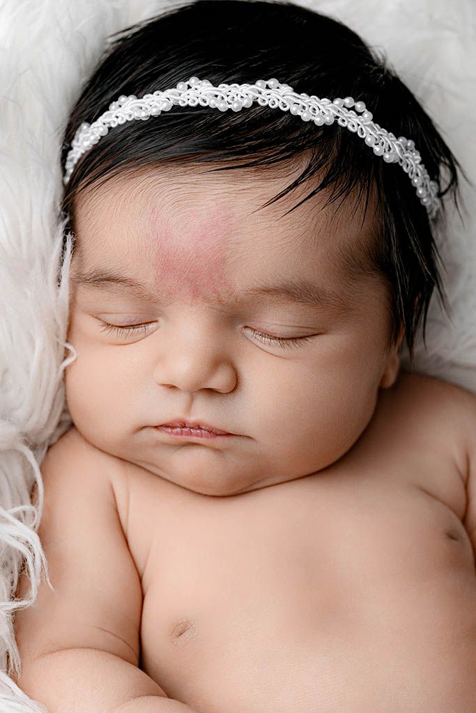 Close-up portrait of sleeping newborn baby girl with dark hair wearing a white lace headband.