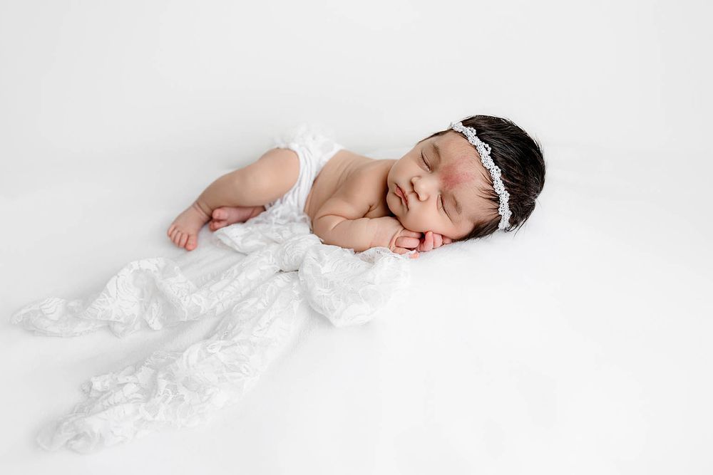 Peaceful newborn baby girl sleeping in white lace wrap and headband on white backdrop.