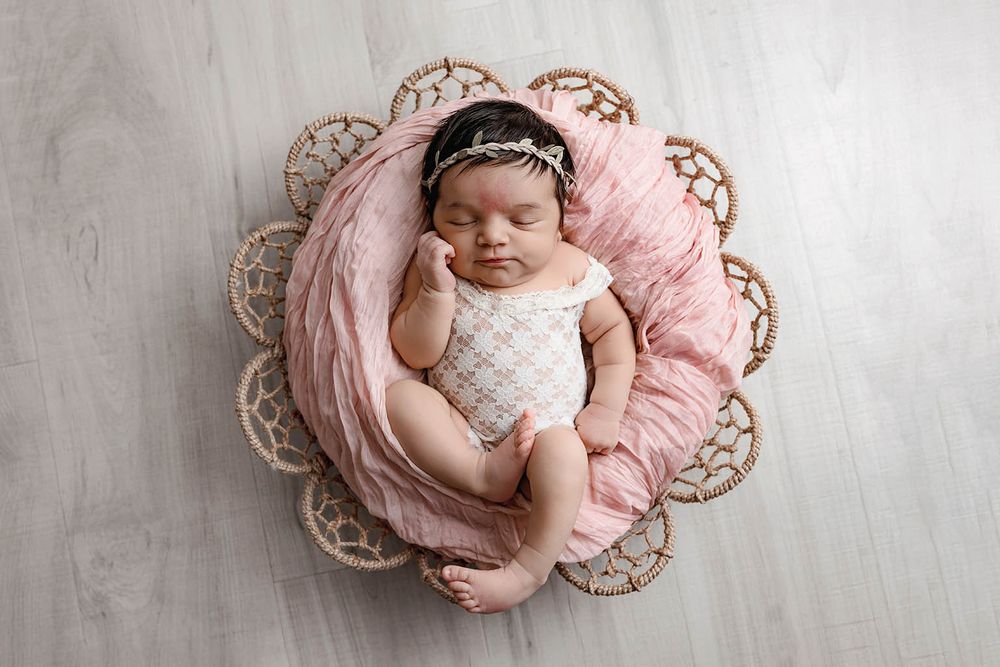 Peaceful newborn baby girl in white romper with floral headband resting in pink-lined basket.