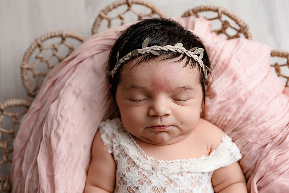 Peaceful newborn baby girl in lace outfit with leaf headband resting in wicker basket with pink fabric.