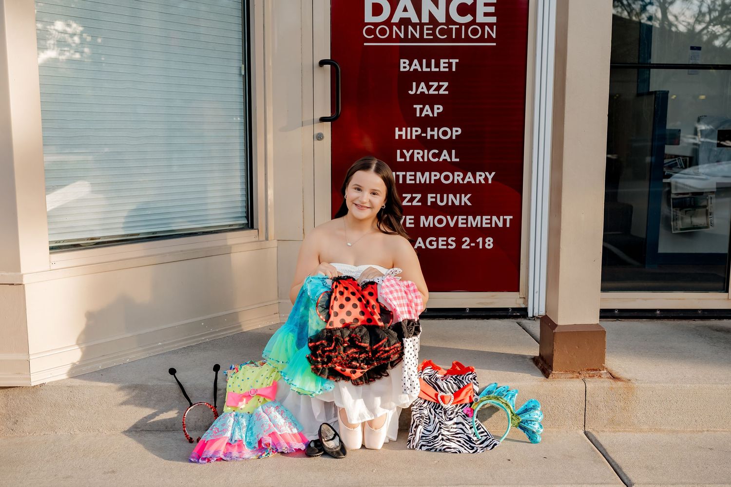 Young dancer sits outside Dance Connection studio holding colorful costumes, with dance styles listed on window.