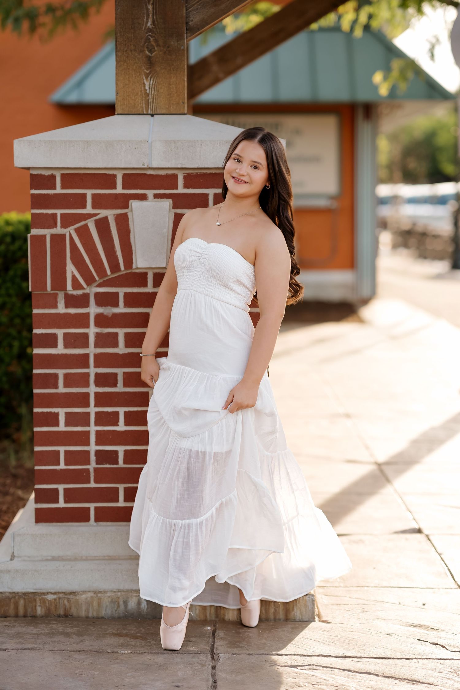Young woman in white strapless maxi dress and ballet flats poses gracefully beside a brick column on a sidewalk.