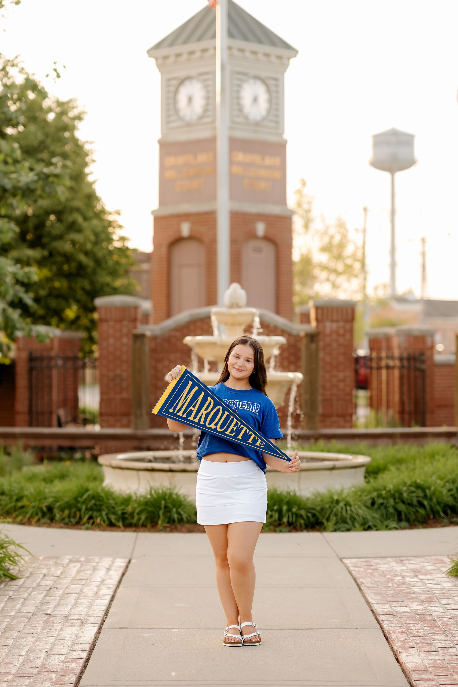 Young woman holding a Marquette University pennant flag in front of a brick clock tower and fountain.