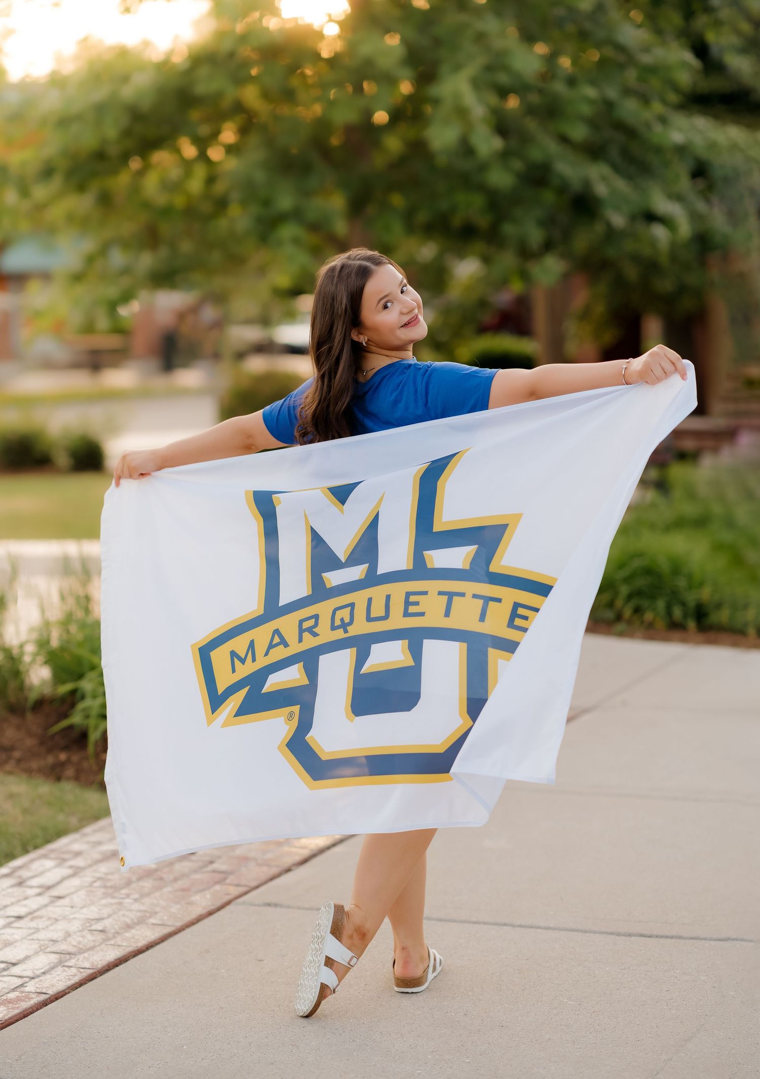 Young woman holding a large white Marquette University flag outdoors, wearing a blue shirt and white sandals.