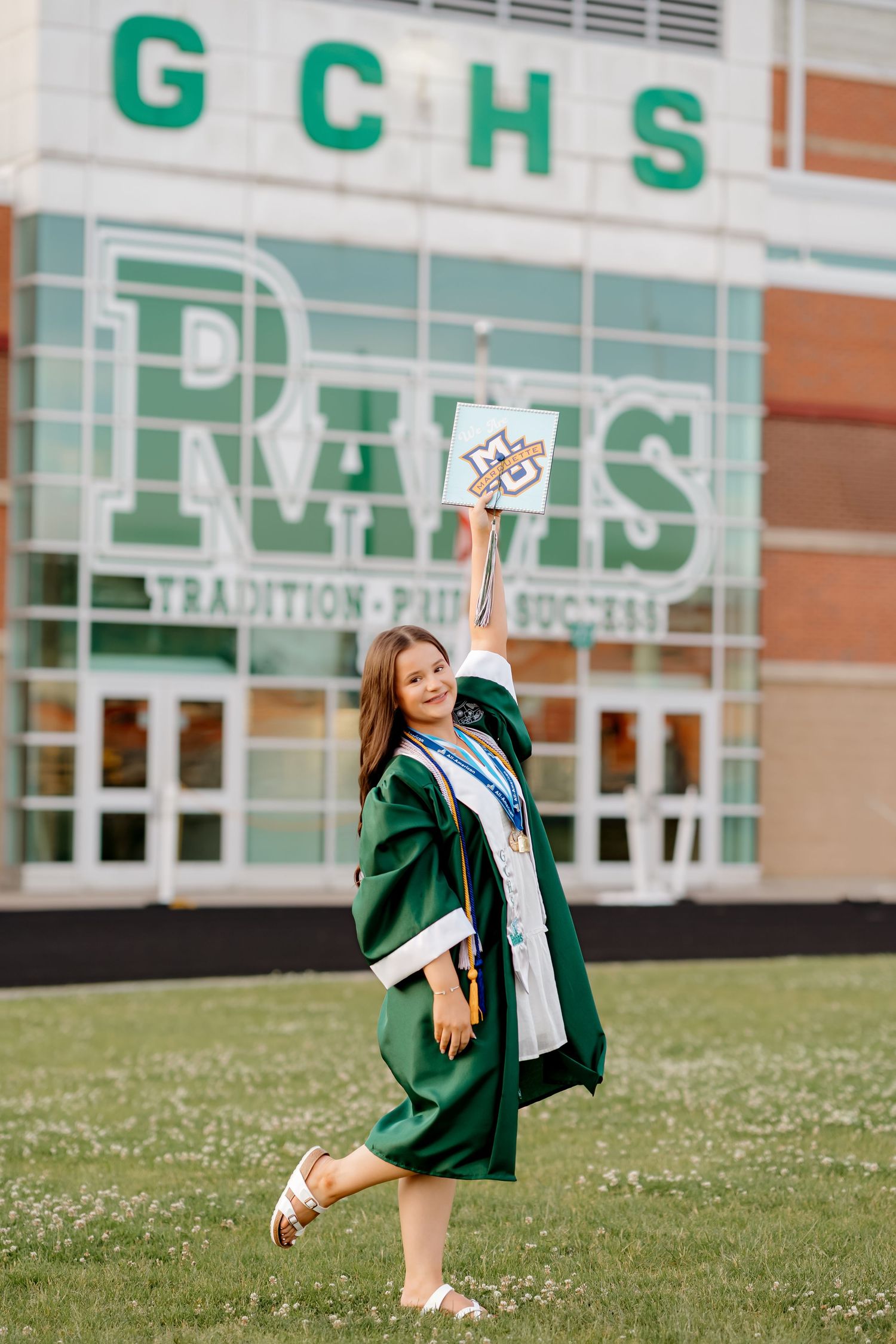 High school graduate in green cap and gown holds diploma aloft outside GCHS Rams school building.