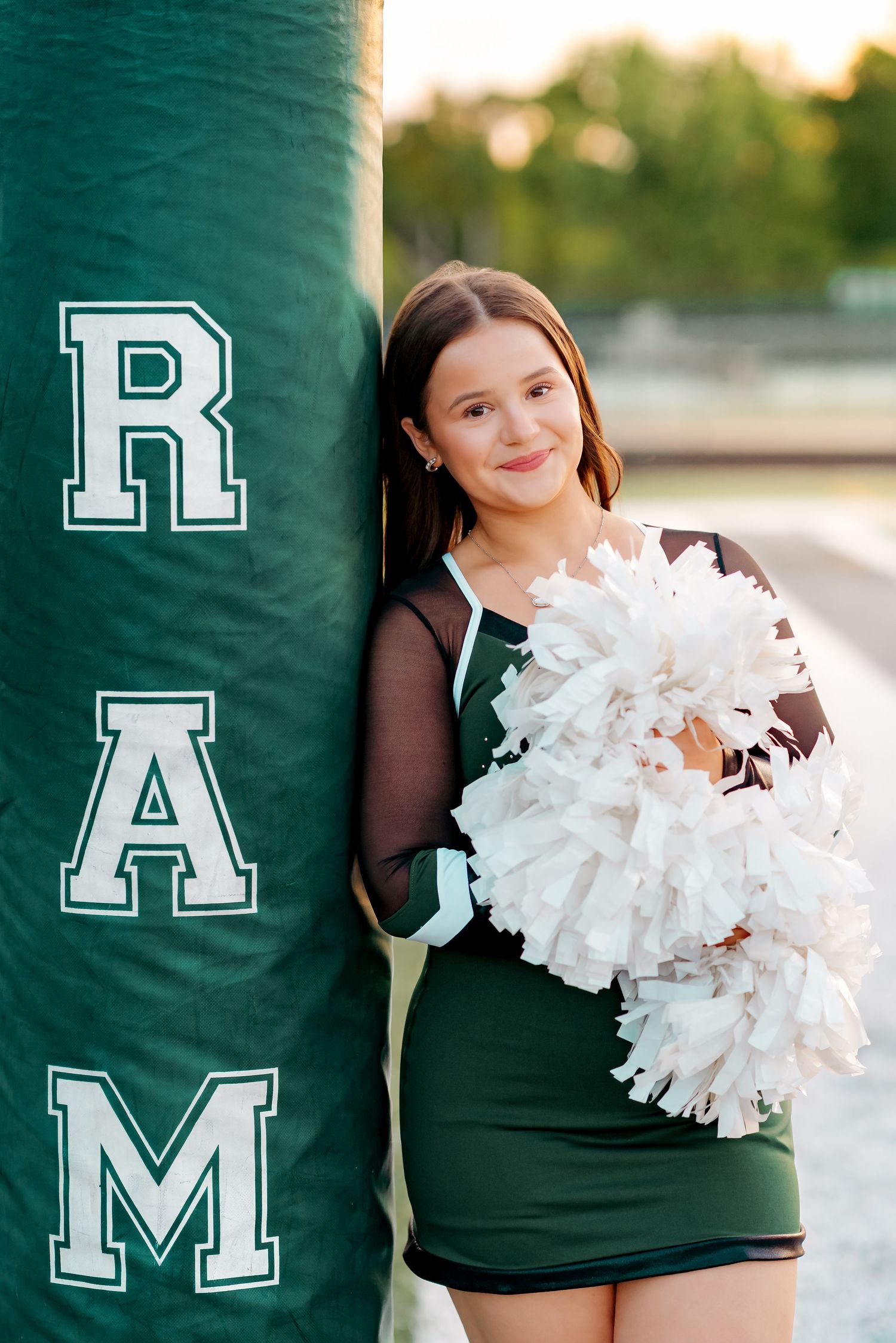 Young cheerleader in green uniform holds white pom-poms, leaning against a green RAM mascot padding on a field.