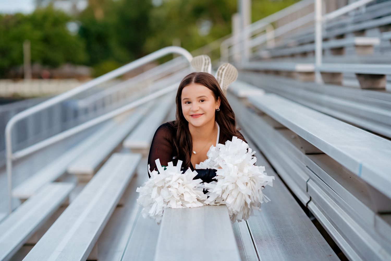 Young cheerleader with pom-poms poses on bleachers wearing a black top and white bow.