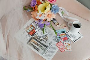 Wedding flat lay featuring colorful paper flower bouquet, magazine, coffee cup, playing cards, and tulle veil on pink chair.
