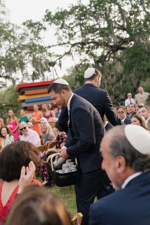 Groomsman collecting broken glass pieces in basket after Jewish wedding ceremony outdoors.