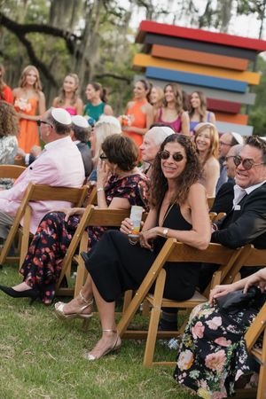 Wedding guests seated in wooden chairs at outdoor ceremony with colorful giant flowers and rainbow sculpture backdrop.