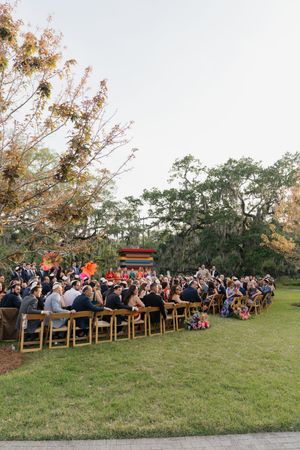 Outdoor wedding ceremony with guests seated on wooden chairs on a green lawn surrounded by trees.