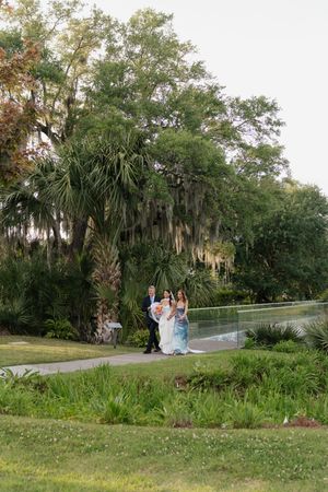 Bride and groom walk along garden path surrounded by Spanish moss-draped trees at dusk.