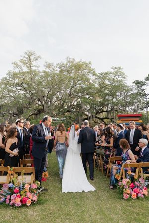 Bride walks down outdoor wedding aisle flanked by colorful floral arrangements and guests seated in wooden chairs under oak trees.