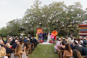 Outdoor Jewish wedding ceremony with guests seated before colorful giant flower chuppah under oak tree.