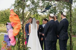 Outdoor Jewish wedding ceremony under floral chuppah with colorful giant paper flowers and guests gathered.