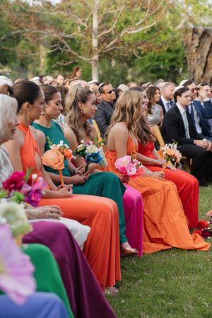 Colorful bridesmaids in vibrant orange, green, and pink gowns seated at an outdoor wedding ceremony.