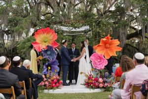 Bride and groom exchange vows under giant paper flowers at outdoor Jewish wedding ceremony with guests.
