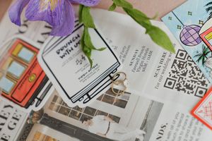 Flat lay of a book-shaped sticker on newspaper with purple iris flowers and colorful cards.