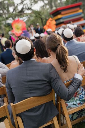 Wedding guests wearing kippot sit together watching outdoor Jewish wedding ceremony under oak trees.