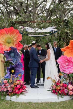 Bride and groom exchange vows under floral chuppah with oversized colorful flowers at outdoor Jewish wedding ceremony.