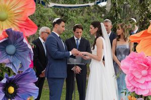 Groom places ring on bride's finger under colorful giant flower wedding arch outdoors.