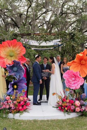 Couple exchanges vows under colorful giant paper flower arch at outdoor Jewish wedding ceremony.
