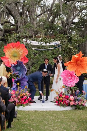 Groom stomps glass at outdoor Jewish wedding ceremony with giant floral decorations under oak trees.