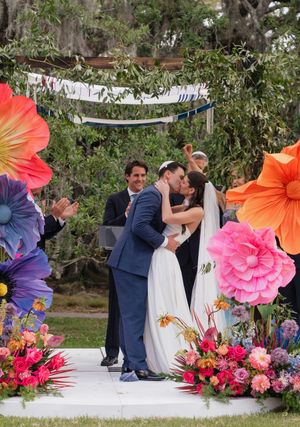 Couple shares first kiss at colorful outdoor Jewish wedding ceremony with giant paper flowers.